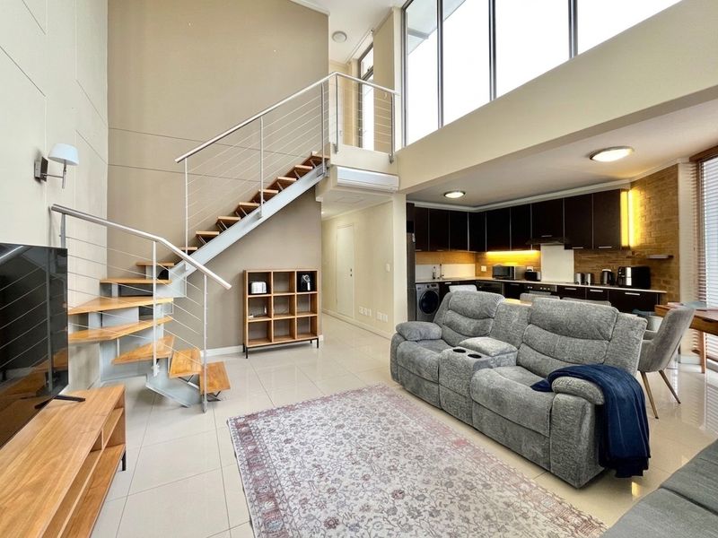 Spacious open-plan living area featuring a modern staircase, high ceilings with large windows, and a well-appointed kitchen with dark cabinetry.