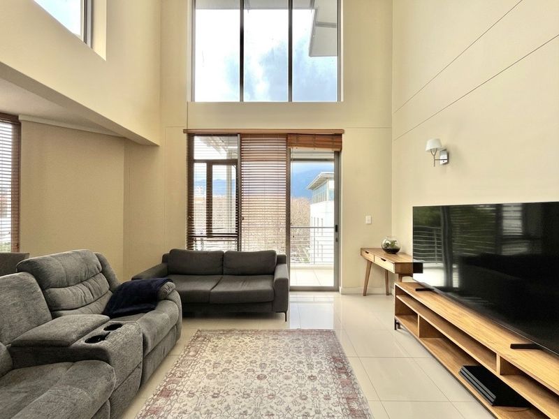 Spacious living room featuring double-volume ceilings, large windows for natural light, and direct access to a balcony with tiled flooring.