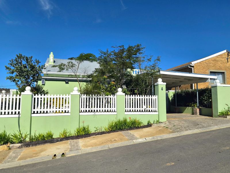 Front elevation of a house featuring a green masonry boundary wall, white picket fence, and a covered carport with a paved driveway.