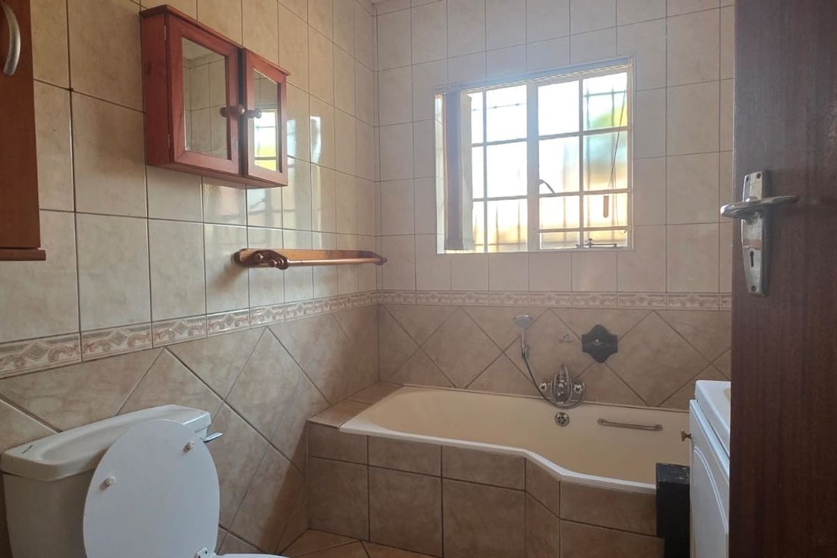 A functional bathroom featuring tiled walls and flooring, a built-in bathtub, a toilet, and a window with security bars.