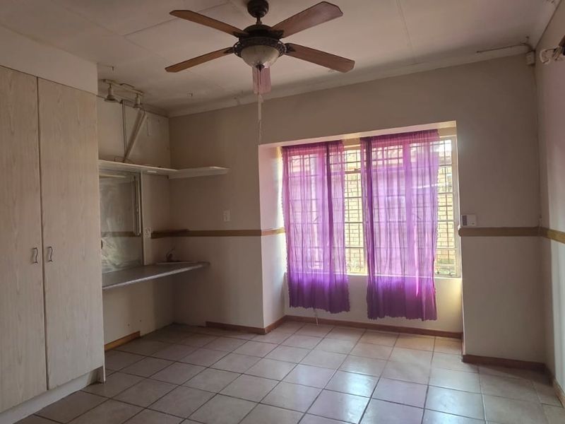 A spacious bedroom featuring built-in cupboards, tiled flooring, a ceiling fan, and large windows with security bars, offering a blank canvas for renovation.