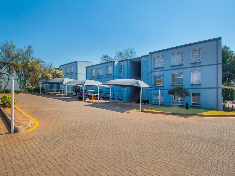 Exterior view of a multi-story apartment complex featuring a paved driveway and dedicated shade cloth carport parking bays.