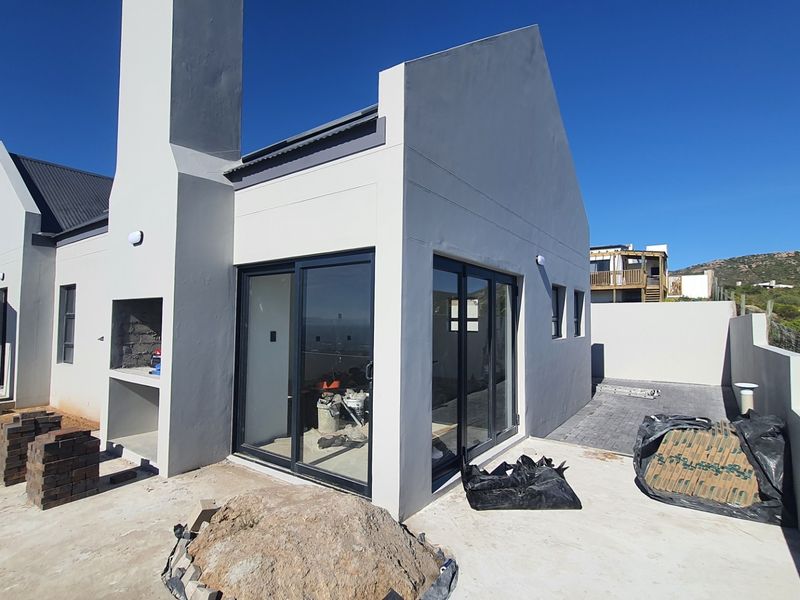 Rear exterior view of a modern house under construction, featuring a built-in braai, sliding glass doors, and a paved patio area.