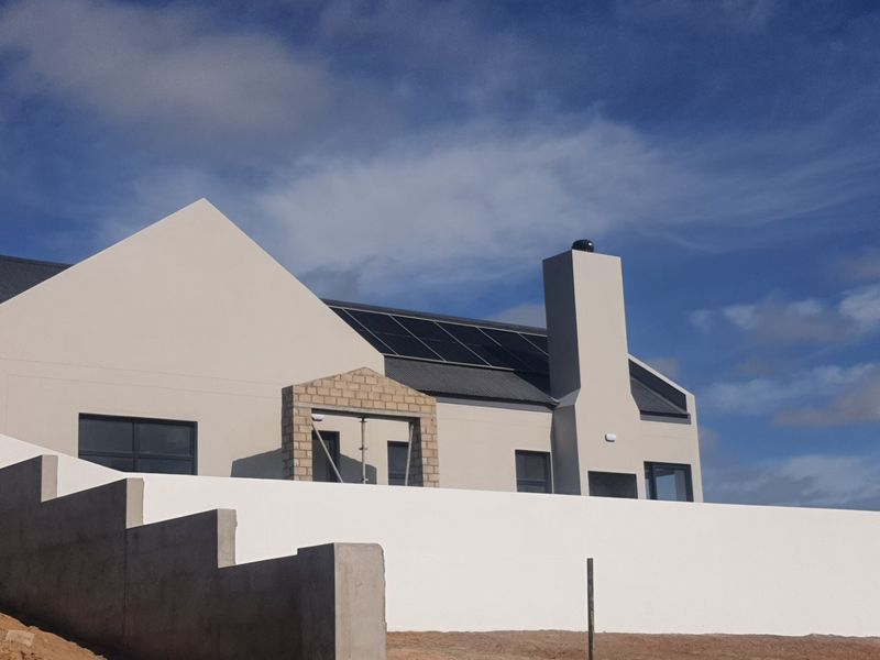 Rear view of a modern house featuring a corrugated metal roof, solar panels, a prominent chimney, and a newly constructed boundary wall.