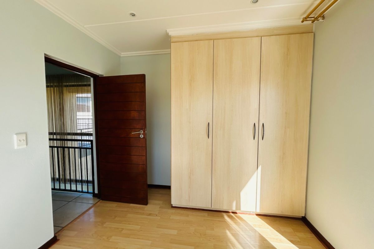 A bright, empty bedroom featuring light wood-look laminate flooring, a built-in wardrobe, and recessed ceiling lighting, with access to an adjacent balcony.