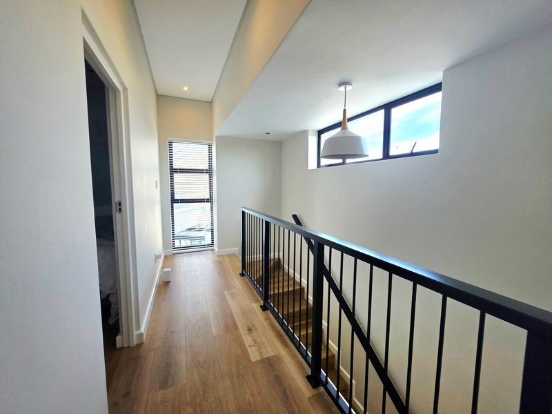 A bright, modern hallway featuring timber flooring, a sleek black metal balustrade, and a staircase with natural light from high-level windows.