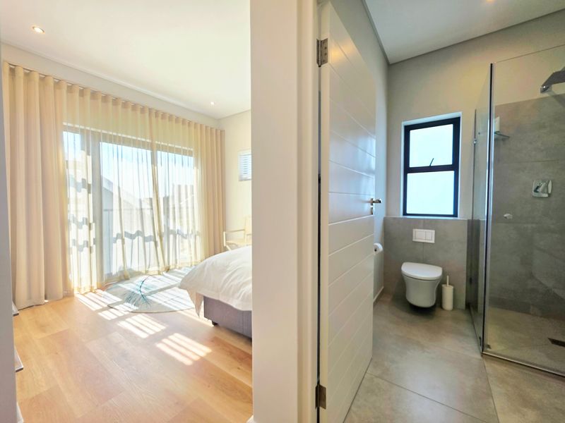 A modern bedroom featuring laminate flooring and sliding glass doors, with an adjacent en-suite bathroom equipped with a glass shower and wall-hung toilet.