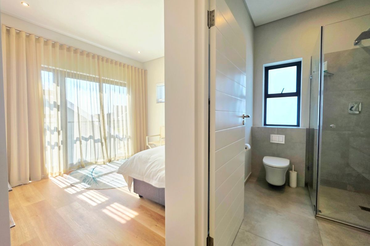 A modern bedroom featuring laminate flooring and sliding glass doors, with an adjacent en-suite bathroom equipped with a glass shower and wall-hung toilet.