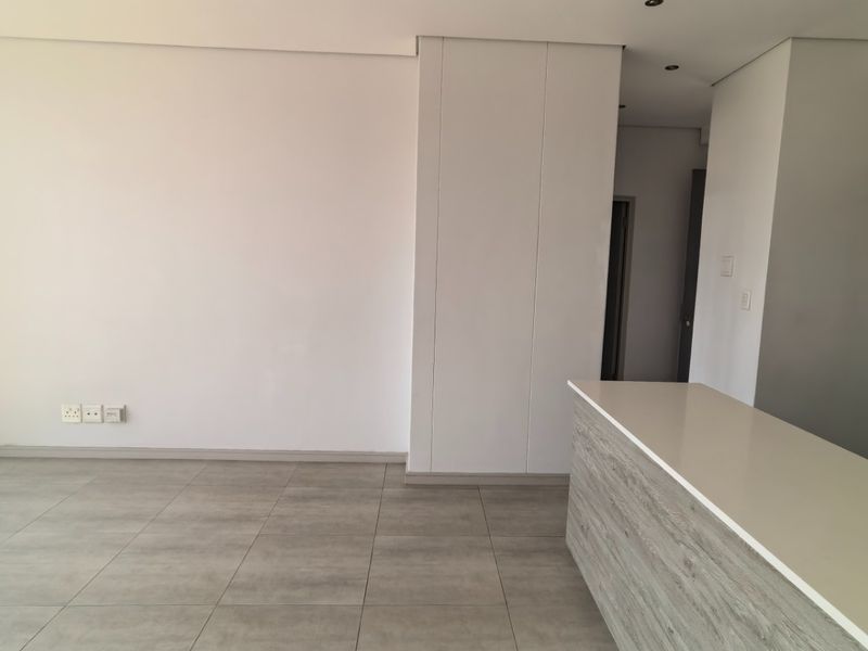 A bright, modern open-plan living and kitchen area featuring neutral tiled flooring, a sleek kitchen island with stone countertop, and floor-to-ceiling glass doors.