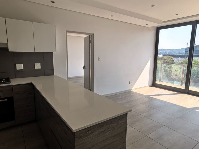 Modern open-plan kitchen and living area featuring sleek cabinetry, stone countertops, integrated appliances, and large sliding doors leading to a balcony with scenic views.