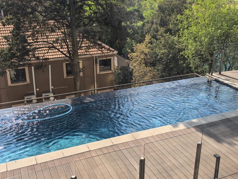 An elevated infinity edge swimming pool featuring modern composite decking and a sleek glass balustrade with stainless steel posts.