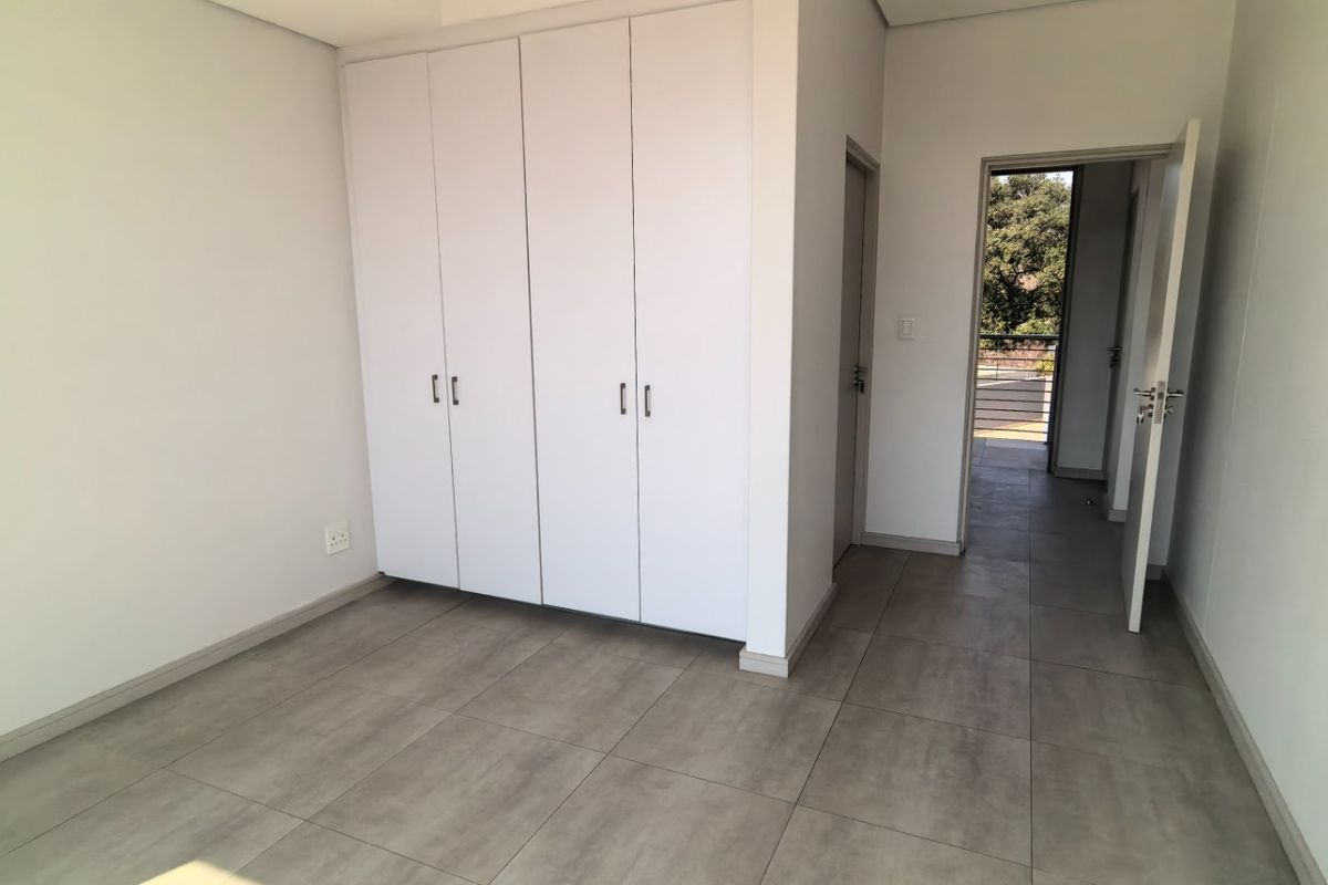 A bright, empty bedroom featuring neutral tiled flooring and ample built-in cupboard storage, leading to an adjacent hallway.