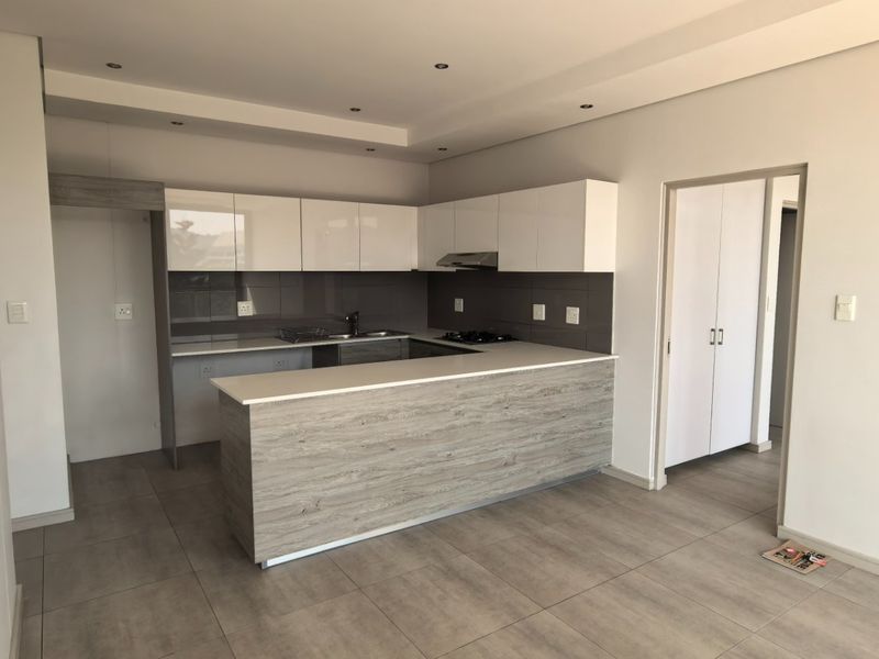 Modern open-plan kitchen featuring sleek white cabinetry, wood-grain breakfast bar, stone countertops, and integrated hob with extractor fan, set on neutral tiled flooring.