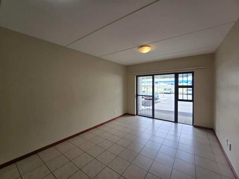 An empty, well-lit living room featuring neutral tiled flooring and a sliding glass door with integrated security gate providing access to the exterior.