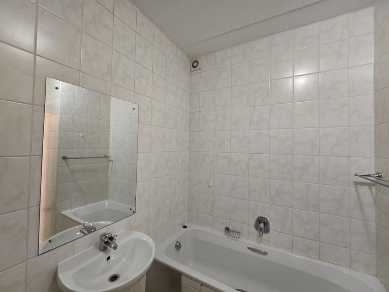 A functional bathroom featuring a bathtub, pedestal basin, and full-height wall tiling, presenting a clean and dated aesthetic ready for modernization.