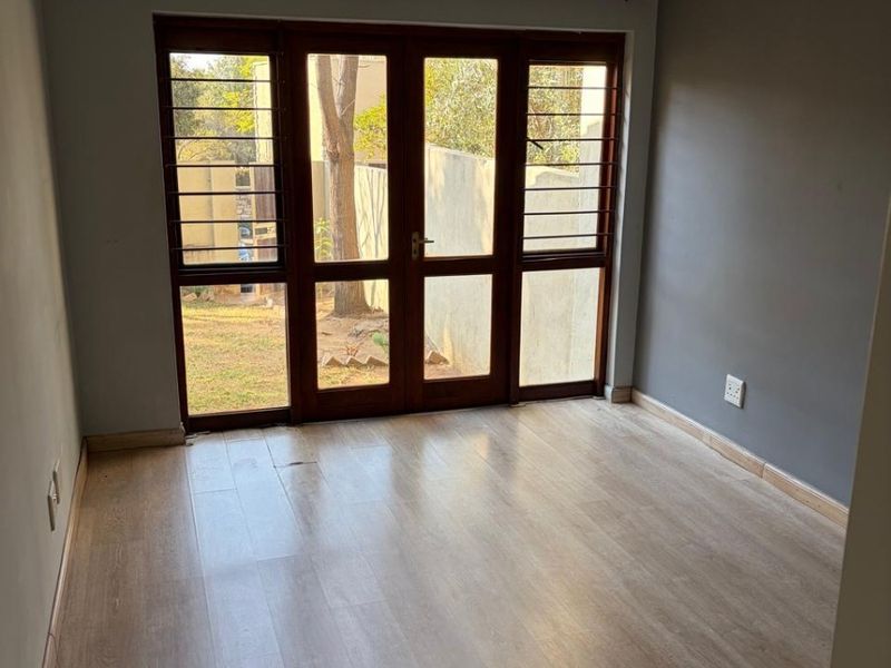 An empty bedroom featuring light-toned laminate flooring, grey walls, and wooden-framed glass doors providing access to the exterior.