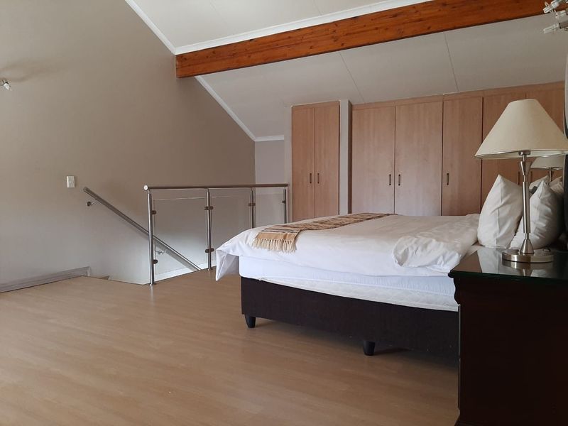 Spacious loft-style bedroom featuring vaulted ceilings, exposed timber beams, built-in wooden cupboards, and a modern glass balustrade overlooking the staircase.