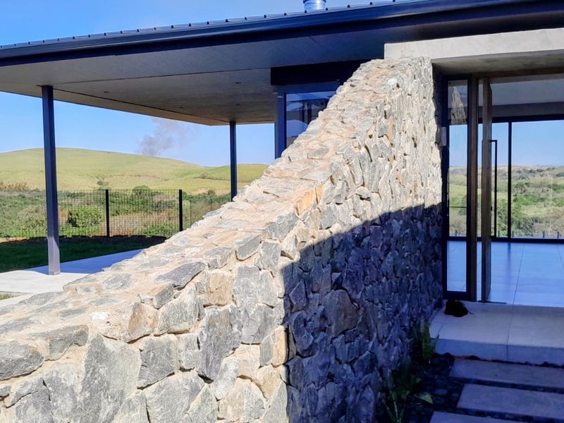 Modern architectural entrance featuring a prominent natural stone feature wall, covered patio area, and expansive glass doors overlooking the surrounding landscape.