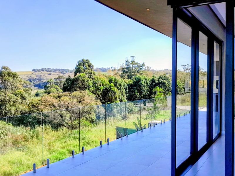 A modern, spacious balcony featuring a frameless glass balustrade and large sliding doors, offering expansive views of the surrounding landscape.