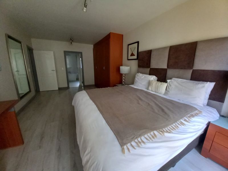 Spacious bedroom featuring laminate flooring, a large built-in wardrobe, and an upholstered headboard, with an adjoining bathroom visible through the doorway.