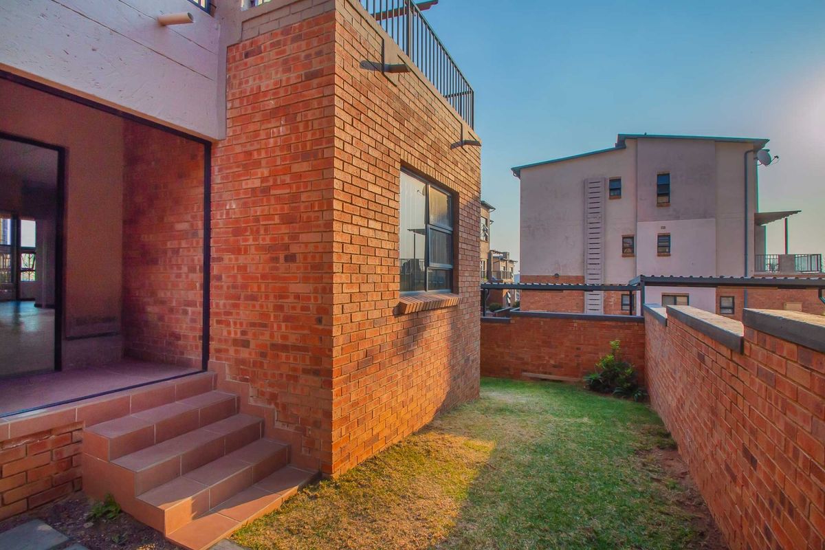 A rear exterior view of the apartment featuring exposed brickwork, a small private lawn, and tiled steps leading to the interior living space.