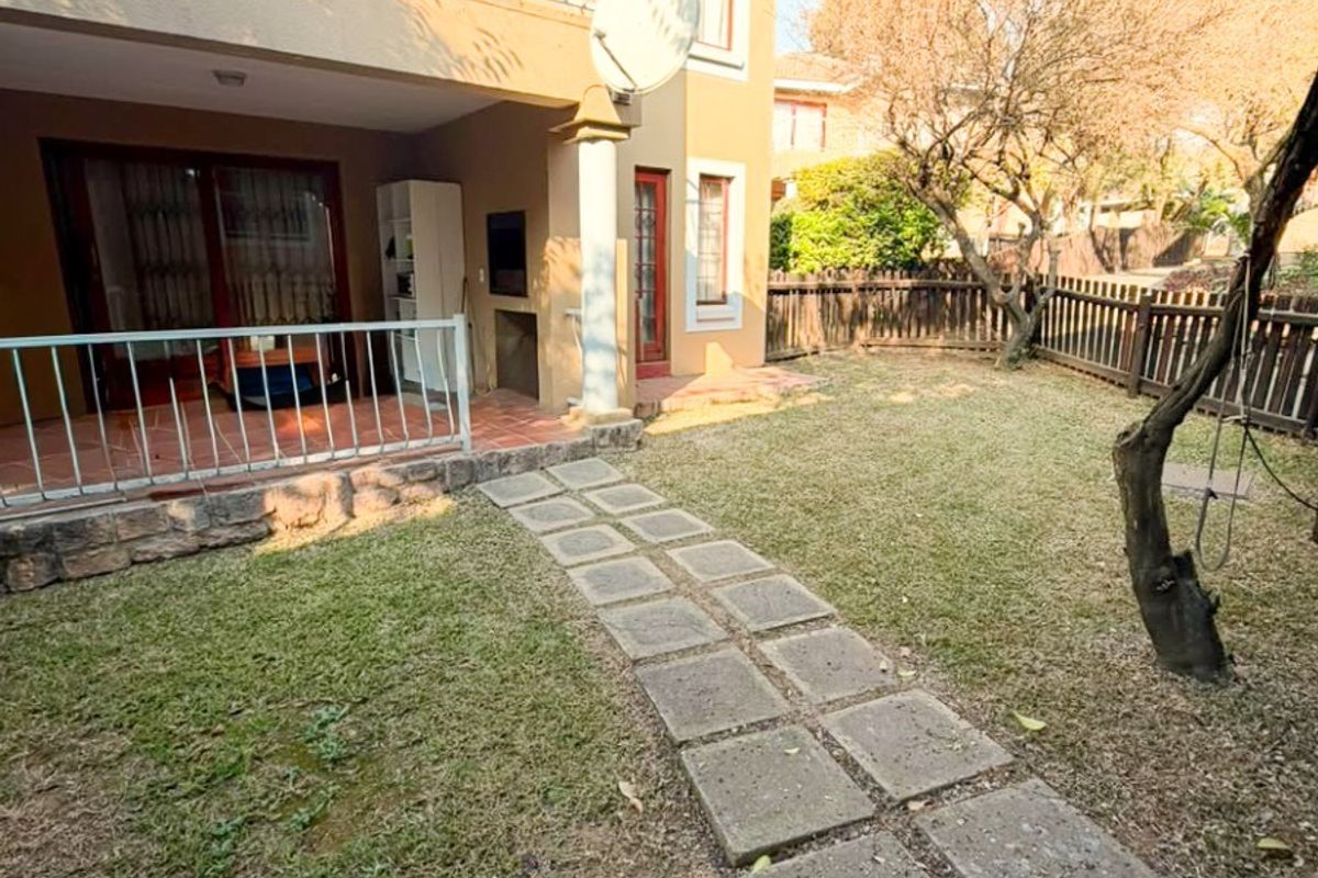 Rear exterior view of the townhouse featuring a covered patio with a built-in braai, a paved garden path, and an upper-level balcony.