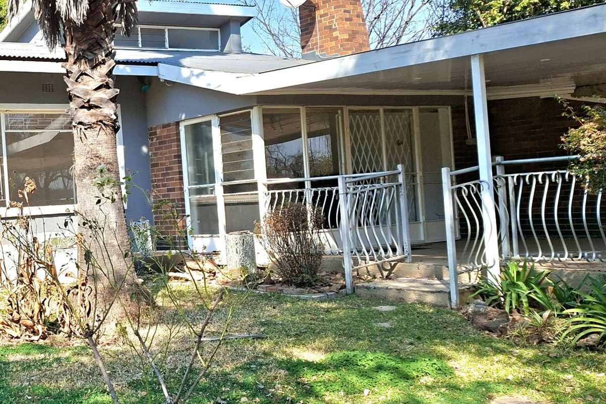 Rear view of the house featuring a covered patio area, brick chimney, and metal security features, set within a garden space.