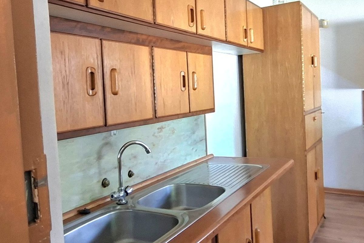 A dated kitchen area featuring original wooden cabinetry, a double stainless steel sink, and laminate countertops, offering potential for a modern renovation.