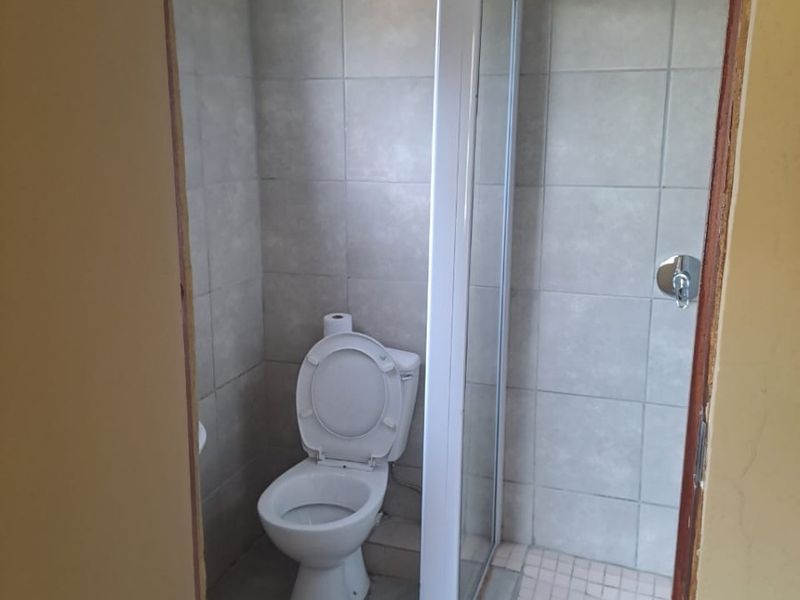 A compact bathroom featuring a shower enclosure and toilet with neutral wall and floor tiling, currently requiring finishing touches to the door frame.