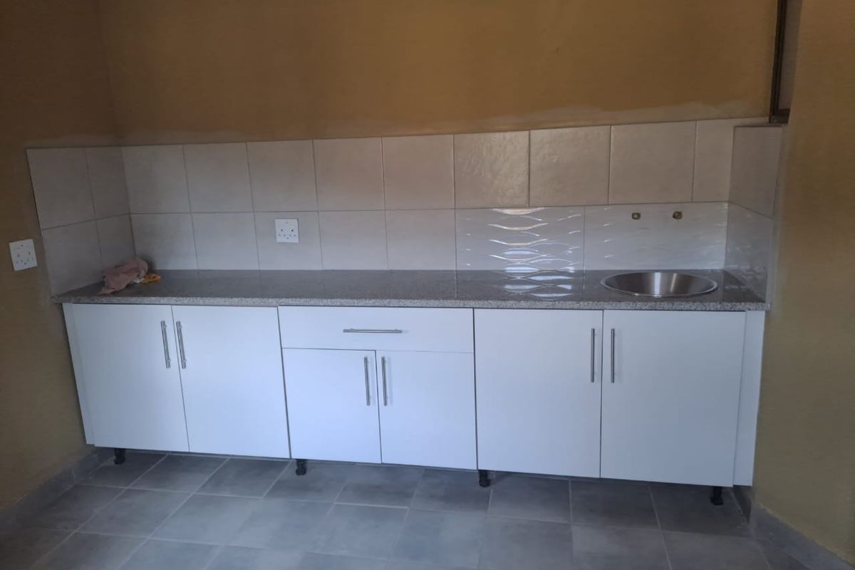 A functional kitchen area featuring white built-in cabinetry, granite countertops, a stainless steel sink, and tiled backsplash, currently awaiting final finishing touches.