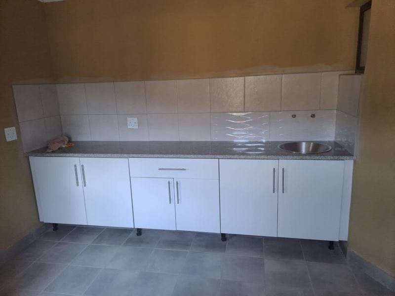 A functional kitchen area featuring white built-in cabinetry, granite countertops, a stainless steel sink, and tiled backsplash, currently awaiting final finishing touches.