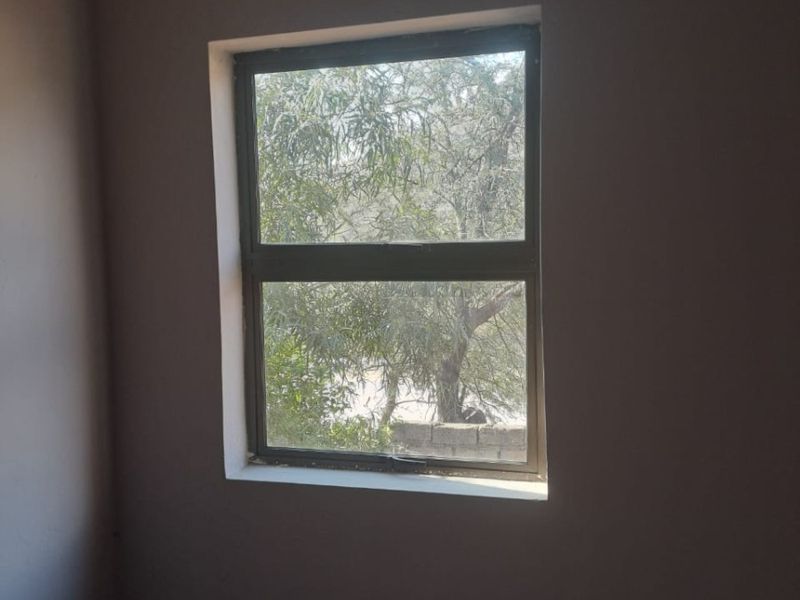 A simple, empty bedroom space featuring a neutral wall finish and a standard aluminium window frame looking out onto greenery.