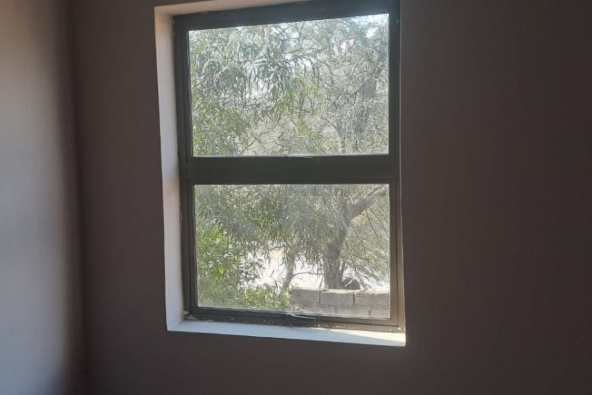 A simple, empty bedroom space featuring a neutral wall finish and a standard aluminium window frame looking out onto greenery.