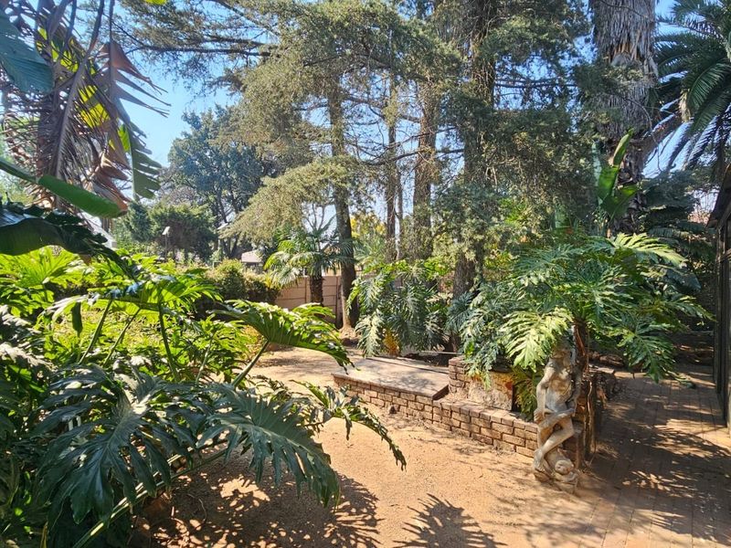 A lush, mature garden featuring established trees, a brick retaining wall, and a paved walkway alongside the property exterior.
