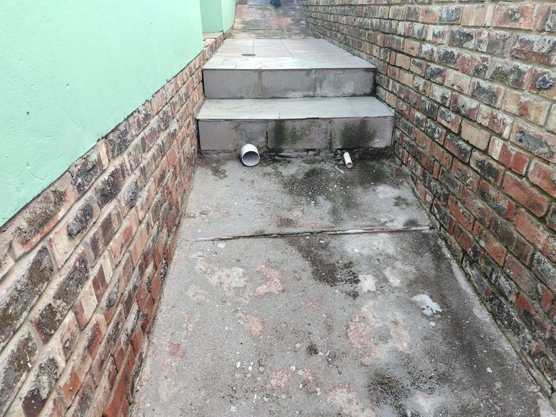 A narrow exterior side passage featuring brick walls, concrete steps, and exposed drainage pipes requiring maintenance and surface cleaning.