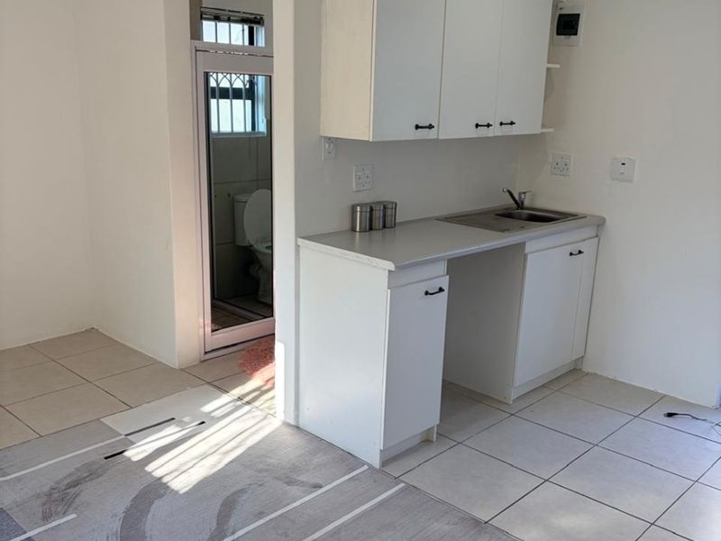 A compact, functional kitchen area featuring white built-in cabinetry, a stainless steel sink, and neutral tiled flooring, with direct access to an adjacent bathroom.
