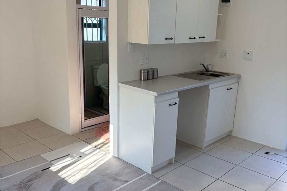 A compact, functional kitchen area featuring white built-in cabinetry, a stainless steel sink, and neutral tiled flooring, with direct access to an adjacent bathroom.