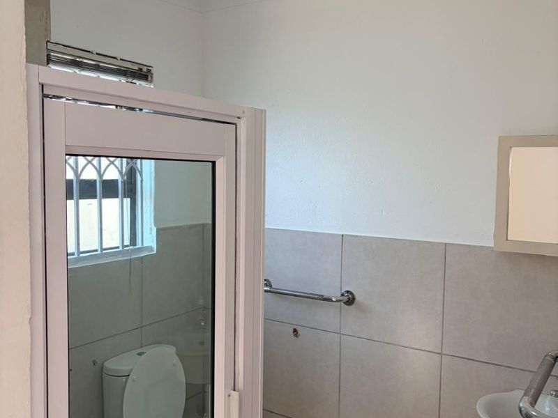 A functional bathroom featuring tiled walls, a wall-mounted basin, a toilet, and a safety grab rail, with a window fitted with security bars.