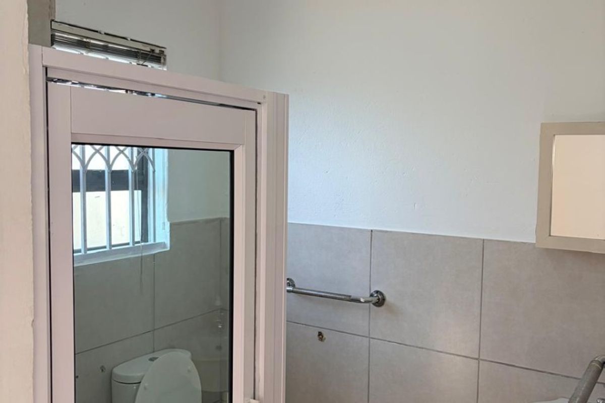 A functional bathroom featuring tiled walls, a wall-mounted basin, a toilet, and a safety grab rail, with a window fitted with security bars.