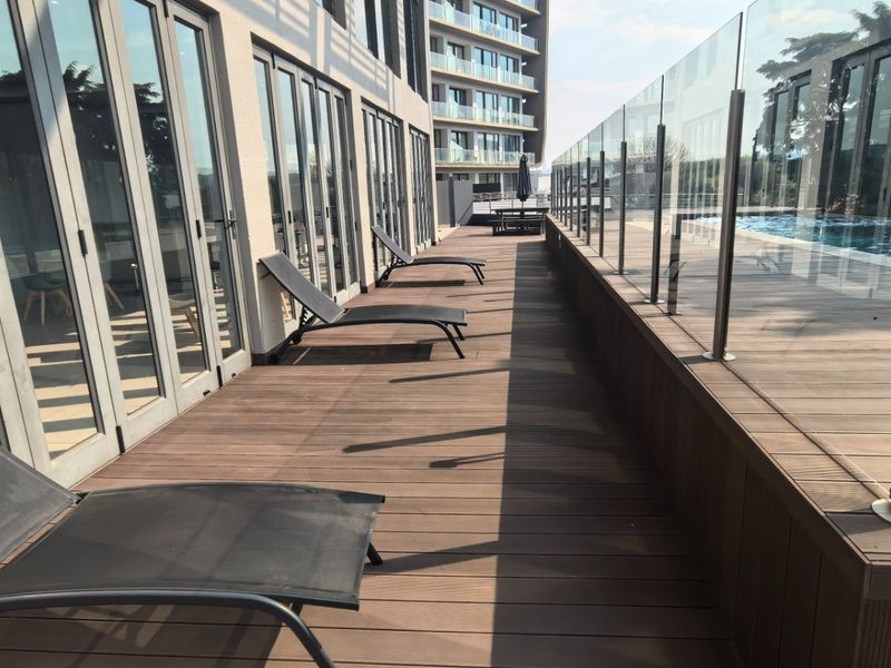 A modern pool deck area featuring timber flooring, glass balustrades, and direct access to the apartment interior through large bi-fold doors.
