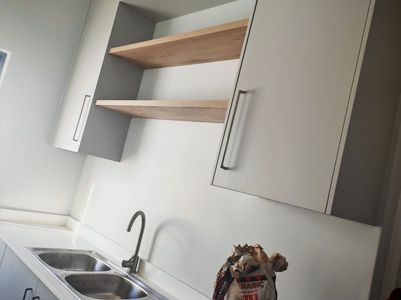 A modern kitchen area featuring light grey cabinetry, open timber shelving, a double bowl stainless steel sink, and a contemporary gooseneck mixer tap.
