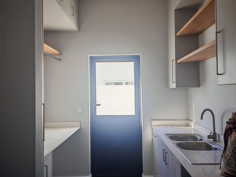 A modern, well-appointed scullery featuring sleek cabinetry, stone countertops, a double sink with a gooseneck tap, and open shelving for organized storage.