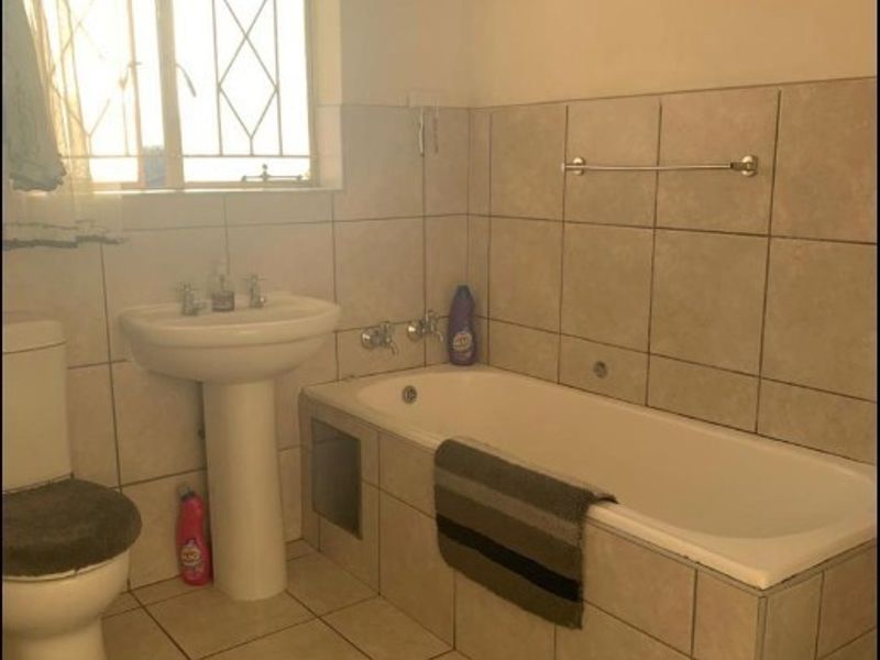 A functional bathroom featuring a pedestal washbasin, a built-in bathtub with tiled surround, and neutral ceramic wall and floor tiling.
