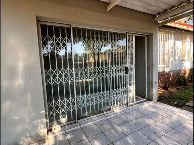 A covered outdoor patio area featuring tiled flooring, a sliding glass door with a security gate, and a simple overhead roof structure.