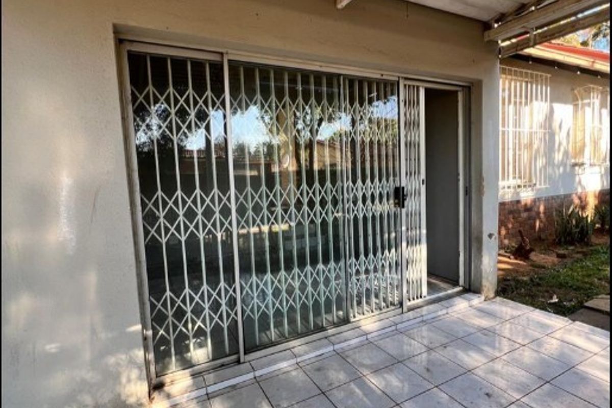 A covered outdoor patio area featuring tiled flooring, a sliding glass door with a security gate, and a simple overhead roof structure.