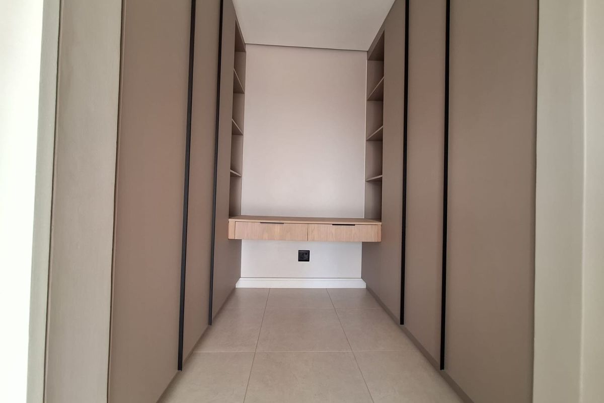 A modern walk-in closet or dressing area featuring floor-to-ceiling built-in cabinetry, integrated shelving, and a floating desk with neutral tiled flooring.