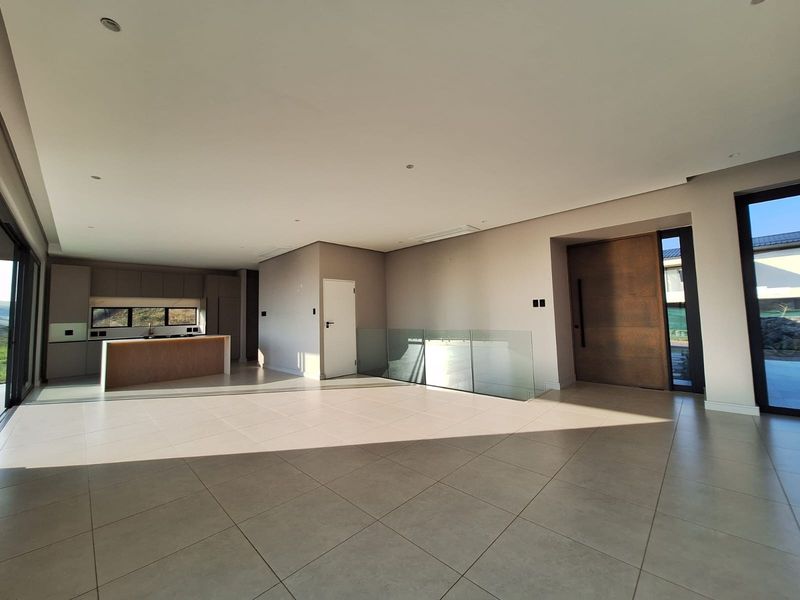 Spacious open-plan living area featuring large format floor tiles, a modern kitchen island, glass balustrade, and a grand timber pivot entrance door.