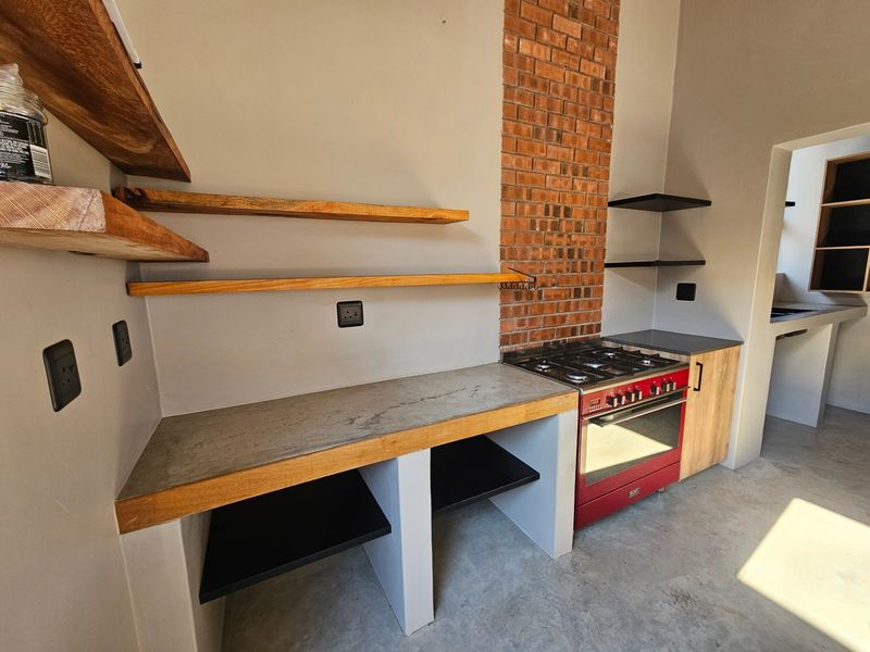 Modern industrial-style kitchen featuring exposed brickwork, concrete countertops, open timber shelving, and a vibrant red built-in gas stove.