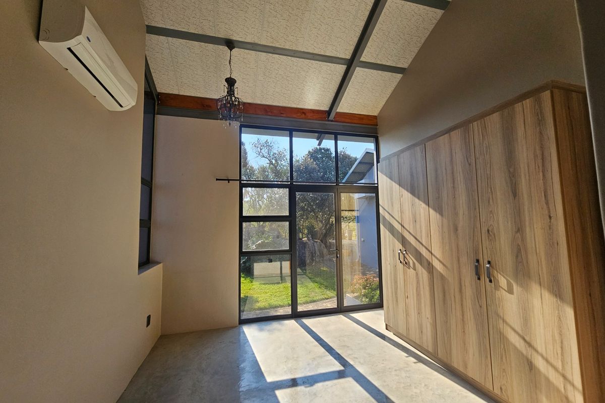 A bright, modern bedroom featuring polished concrete floors, built-in wooden cupboards, exposed ceiling beams, and large glass doors opening to the garden.