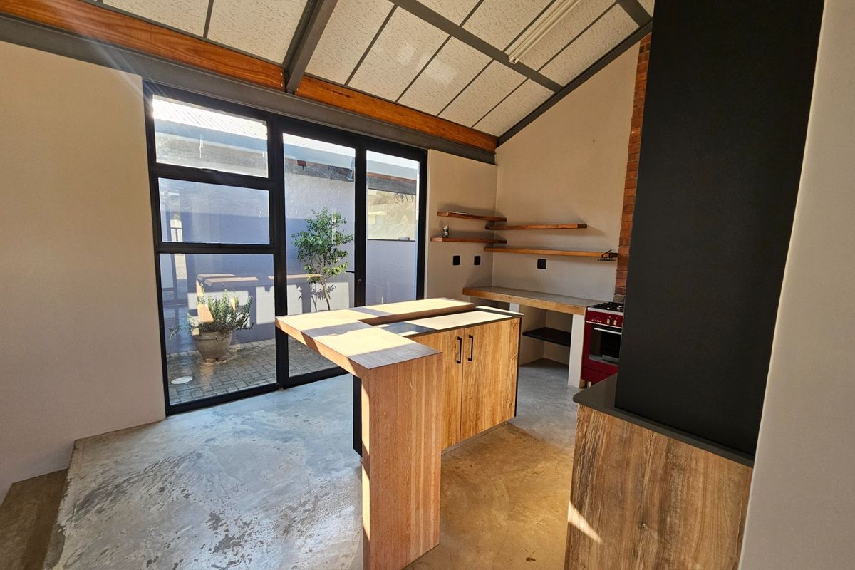 Modern industrial-style kitchen featuring a central island, exposed timber beams, concrete flooring, and large glass doors opening to an outdoor area.